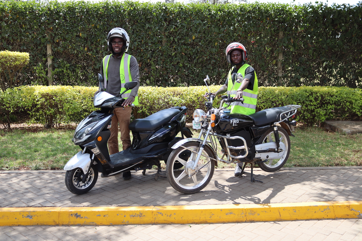 Two men on a sidewalk wearing helmets; each is standing next to an electric bike